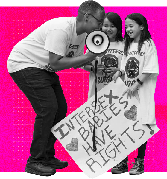 Saifa, holding a megaphone, crouches over two intersex kids at a protest. All are wearing End Intersex Surgery t-shirts and smiling. The kids hold a sign saying 'Intersex Babies Have Rights' painted with hearts.